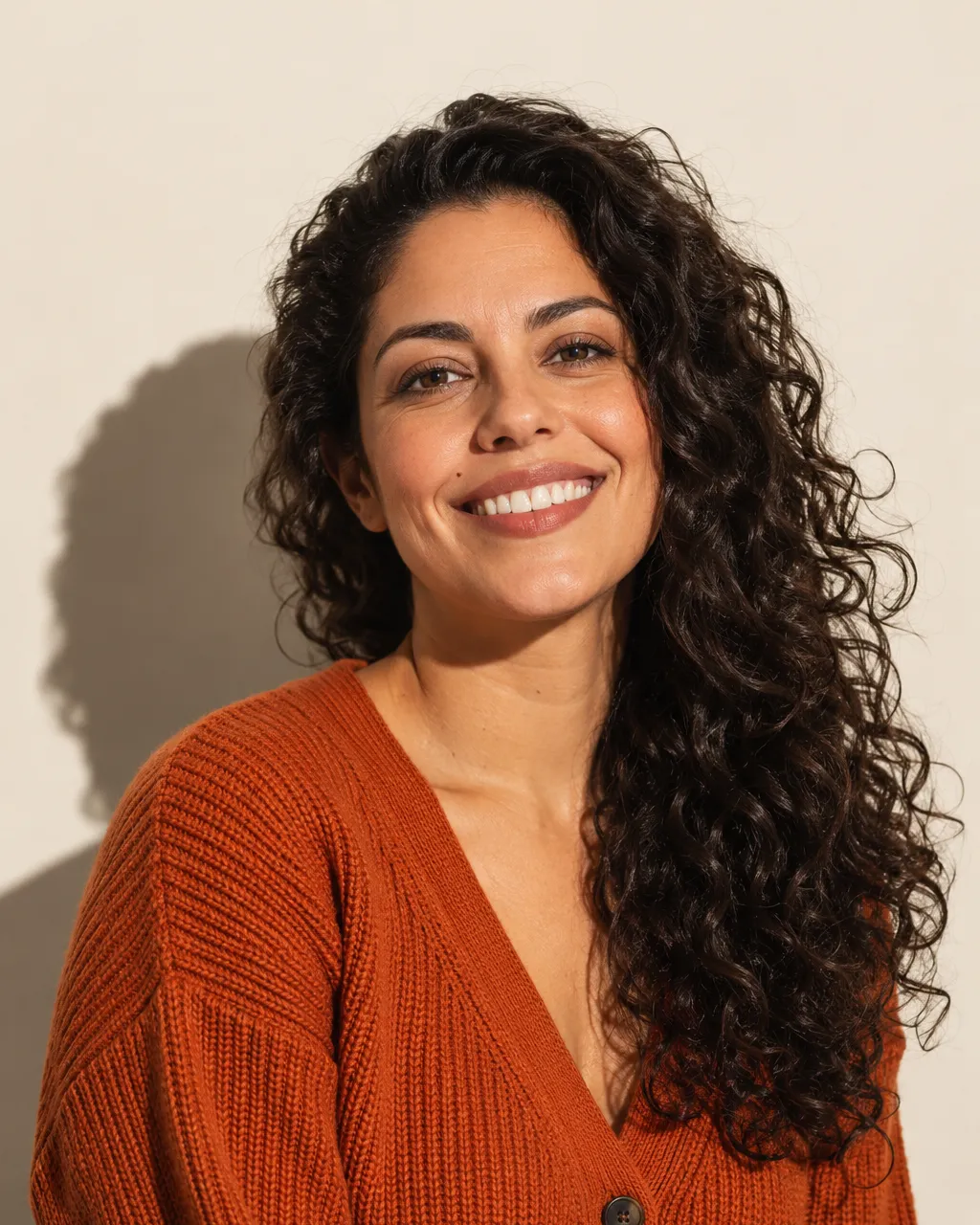 Studio portrait of a Brazilian-Portuguese woman in her mid-thirties with long dark curly hair, wearing a rust-orange…