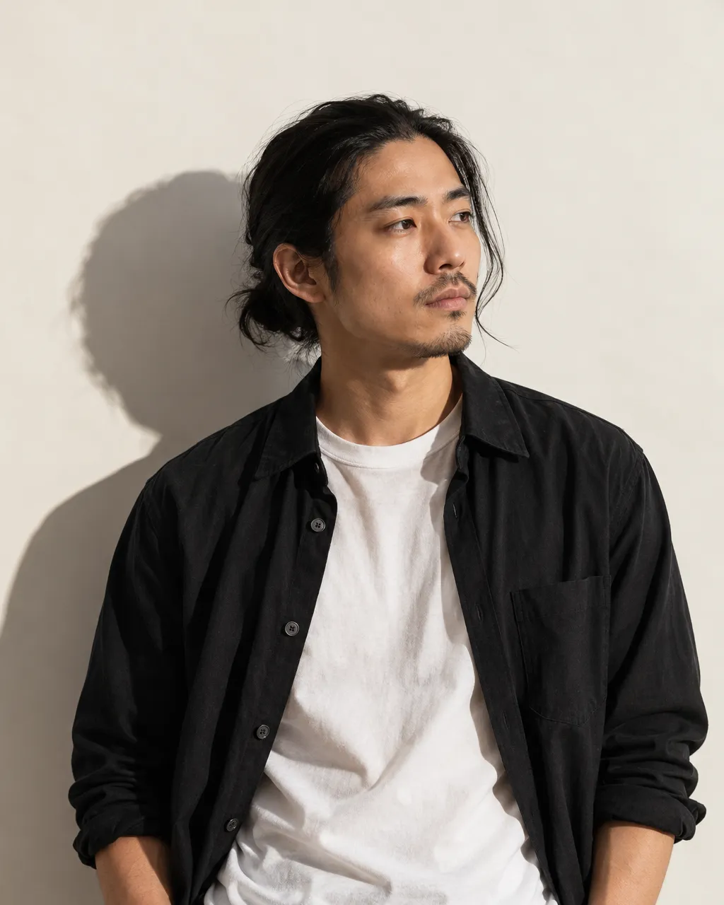 Studio portrait of a Japanese man in his early thirties with shoulder-length straight black hair tied back, wearing…