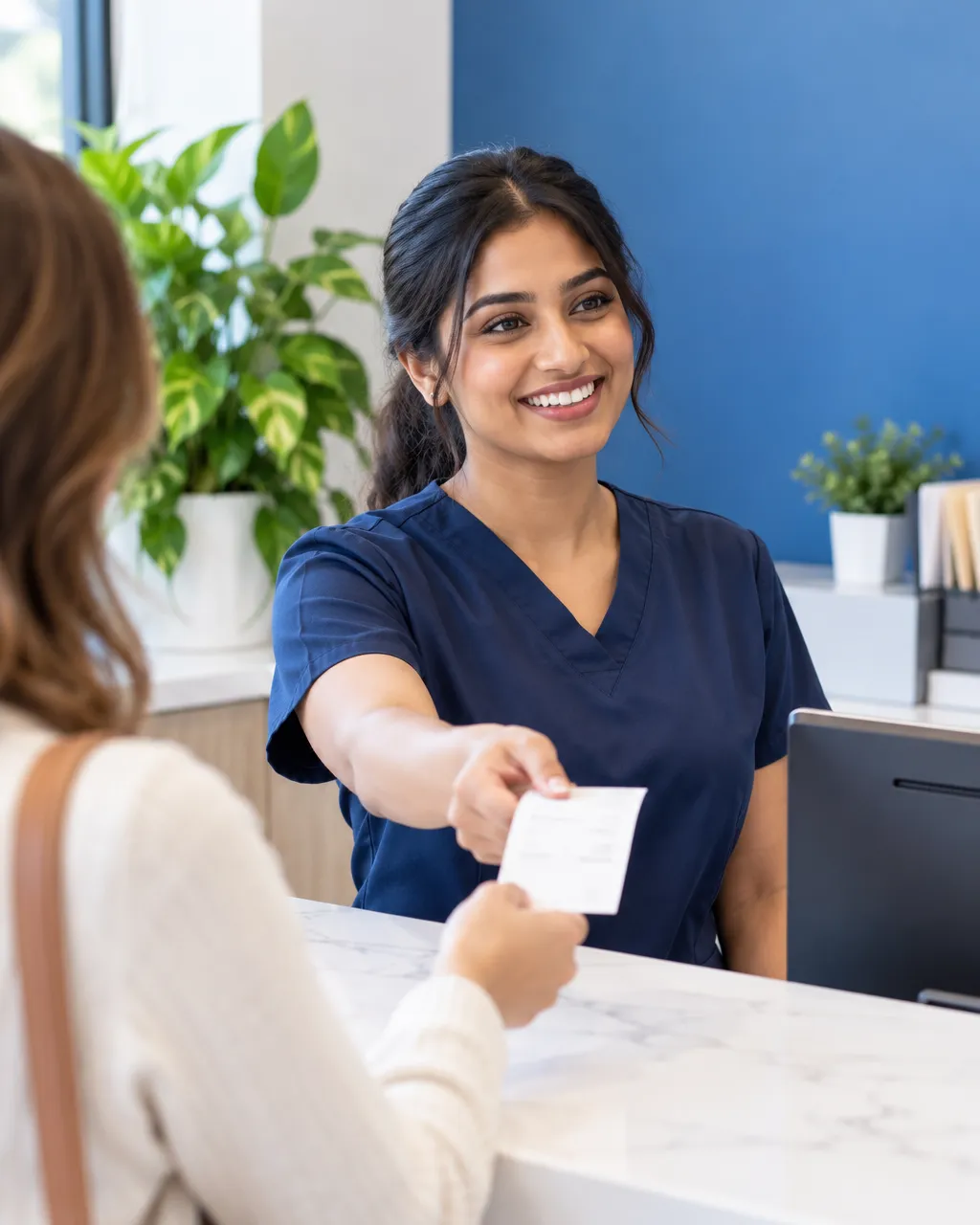 Friendly front-desk team member, a young South Asian woman in navy scrubs, smiling as she hands a printed receipt to…