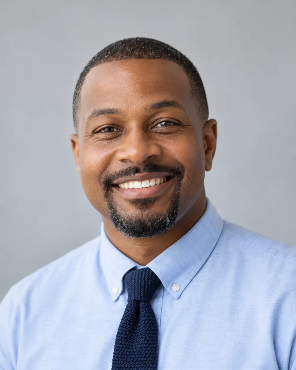 Professional portrait of a Black man in his forties, lower school director, close-cropped hair, light blue oxford…